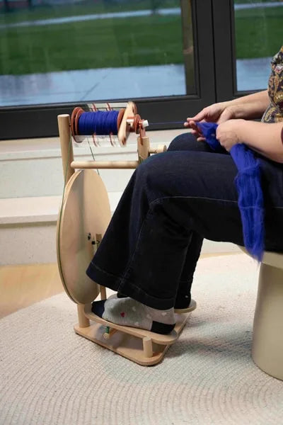 Person spinnt mit dem Lojan Buddy Doppeltritt-Spinnrad, blaues Garn auf der Spule, ergonomisches Arbeiten im Sitzen mit modernem Holzspinnrad